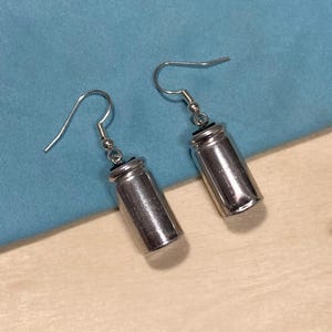 May include: A pair of silver-colored earrings. Each earring features a cylindrical design with a small, dark cap and a curved hook for the ear. The earrings are displayed against a light blue background.