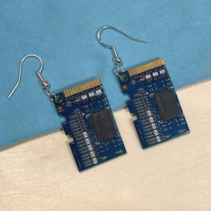 May include: A pair of unique earrings made from blue circuit board material. The earrings have a rectangular shape with gold-colored edges and silver-colored ear wires. The circuit boards have intricate patterns and dark gray components.