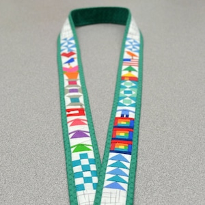 May include: A green lanyard with a white background and colorful patchwork designs. The lanyard has a silver key ring and the text "Sew Tiny" and "A Mini Paper-Pieced Lanyard by Deonn Stott" is printed on the bottom of the image. The website address "www.quiltscapesqs.com" is printed below the text.