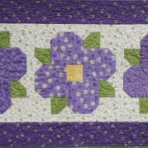Peut inclure: Un chemin de table matelassé violet et vert avec un motif floral. Le chemin de table présente trois grandes fleurs avec des centres jaunes et des feuilles vertes. Le fond est de couleur crème clair avec un motif de pois violets et blancs.