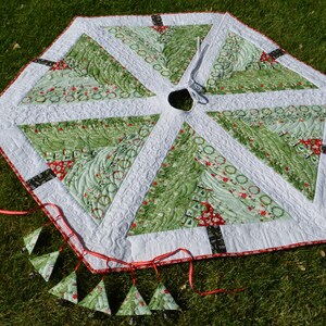 May include: A green and white quilted Christmas tree skirt with a red and white trim. The skirt is decorated with small green and white trees and has a white center with a hole for a tree stand. The skirt is surrounded by a red ribbon with small green and white tree ornaments hanging from it.