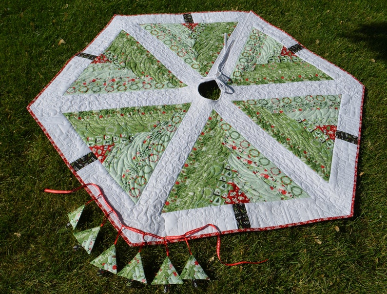 May include: A green and white quilted Christmas tree skirt with a red and white trim. The skirt is decorated with small green and white trees and has a white center with a hole for a tree stand. The skirt is surrounded by a red ribbon with small green and white tree ornaments hanging from it.