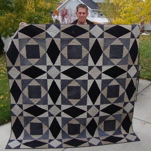 May include: A black, gray, and beige quilt with a geometric pattern. The quilt is folded in half and is being held by a person.
