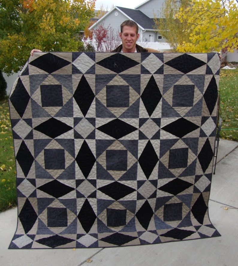 May include: A black, gray, and beige quilt with a geometric pattern. The quilt is folded in half and is being held by a person.