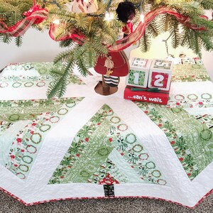 May include: A white quilted Christmas tree skirt with a green and white patterned design. The skirt features a red and white trim and a black and white tree design in the center. The skirt is decorated with a red ribbon and a wooden Christmas countdown block set that reads "...can't wait".