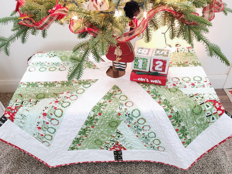 May include: A white quilted Christmas tree skirt with a green and white patterned design. The skirt features a red and white trim and a black and white tree design in the center. The skirt is decorated with a red ribbon and a wooden Christmas countdown block set that reads "...can't wait".
