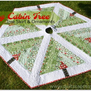 May include: A green and white quilted Christmas tree skirt with a red and black trim. The skirt is shaped like a hexagon and has a hole in the center for a tree stand. The skirt is decorated with a variety of Christmas-themed patterns, including wreaths, trees, and snowflakes. The text "Log Cabin Tree Tree Skirt & Ornaments" is printed on the skirt. The website "www.quiltscapesqs.com" is printed on the bottom of the skirt.
