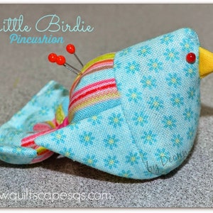Könnte beinhalten: Ein blauer und gelber Vogel-förmiger Nadelkissen mit einem Blumenmuster. Das Nadelkissen hat rote Nadeln, die aus ihm herausragen. Der Text "Little Birdie Pincushion" ist über dem Vogel geschrieben. Der Text "by Deann" ist unter dem Vogel geschrieben. Der Text "www.quiltscapesqs.com" ist unter dem Vogel geschrieben.