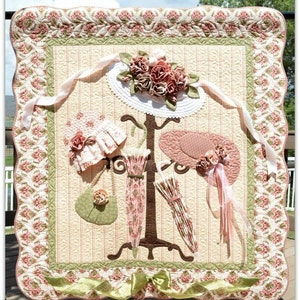 May include: A quilted wall hanging featuring a vintage hat stand with three hats and two umbrellas. The hats are pink and white with floral accents. The umbrellas are pink and white with ruffles. The background is a cream and pink striped fabric with a floral border.