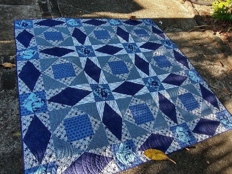 May include: A blue and white quilt with a star pattern. The quilt is made of different fabrics with various blue shades and white accents. The quilt is laid out on a concrete surface.