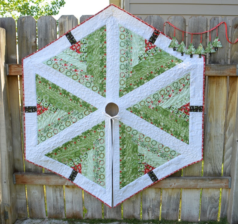 May include: A white hexagonal Christmas tree skirt with a green and red patterned design. The skirt has a white ruffle around the edge and a white tie in the center.