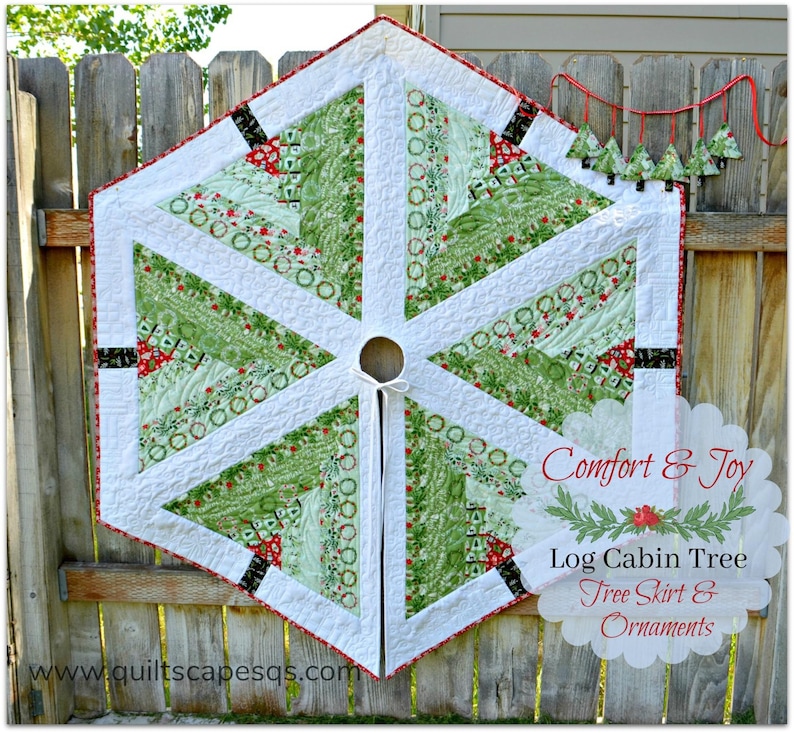 May include: A white, green, and red quilted Christmas tree skirt with a log cabin design. The skirt has a black and white trim and a red ribbon tie. The text "Comfort & Joy Log Cabin Tree Tree Skirt & Ornaments" is printed on a white background.