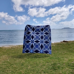 May include: A blue and white quilt with a star pattern is hanging on a clothesline against a backdrop of a beach and ocean. The sky is blue with white clouds.