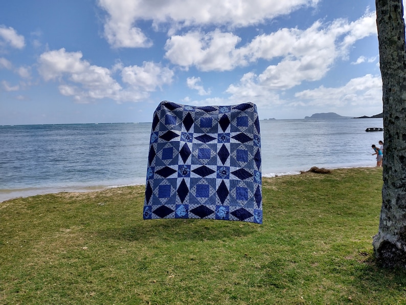 May include: A blue and white quilt with a star pattern is hanging on a clothesline against a backdrop of a beach and ocean. The sky is blue with white clouds.