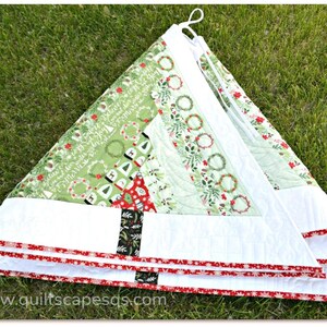 May include: A white quilted Christmas tree skirt with a green and white patterned fabric featuring wreaths and the text "Believe in the magic of Christmas". The skirt has a red and white trim.