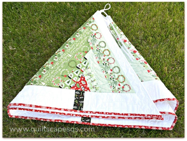 May include: A white quilted Christmas tree skirt with a green and white patterned fabric featuring wreaths and the text "Believe in the magic of Christmas". The skirt has a red and white trim.
