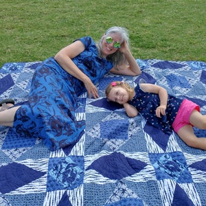 May include: Two people, an adult and a child, are lying on a blue and white patterned quilt on a grassy lawn. The adult is wearing a blue floral dress and sunglasses. The child is wearing a blue and white patterned shirt, pink shorts, and has a pink flower in their hair.