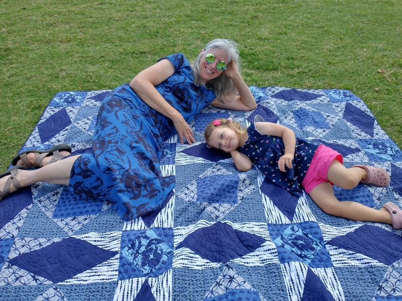 May include: Two people, an adult and a child, are lying on a blue and white patterned quilt on a grassy lawn. The adult is wearing a blue floral dress and sunglasses. The child is wearing a blue and white patterned shirt, pink shorts, and has a pink flower in their hair.