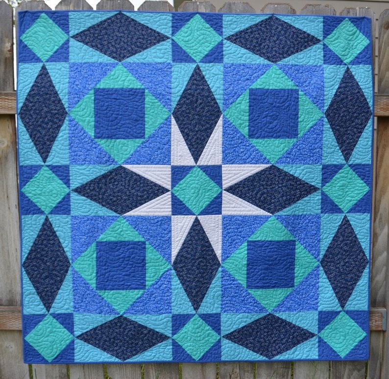 May include: A blue, white, and teal quilt with a starburst pattern in the center. The quilt is made up of squares and diamonds.