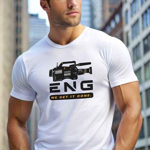 May include: A white short-sleeved t-shirt featuring a black graphic of a professional video camera. Below the camera, the text reads ENG in large bold letters, followed by a yellow rectangular bar with the text WE GET IT DONE.