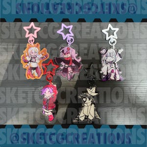 Può includere: Cinque portachiavi in acrilico con personaggi in stile anime, ognuno fissato a una chiusura a forma di stella. I personaggi includono una figura simile a una falena, una persona dai capelli rosa in abito scuro, una figura dai capelli bianchi e una barchetta di carta.