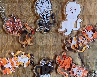 May include: A collection of seven acrylic keychains featuring illustrated big cats in various poses and color schemes, including orange, black, white, and brown patterns. Each charm is attached to a silver-toned metal lobster clasp and ring.