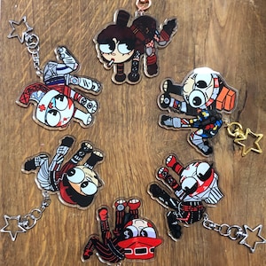 Peut inclure: Six porte-clés de personnages de dessin animé colorés avec des breloques en forme d'étoile. Chaque porte-clés présente un personnage différent avec de grands yeux et un schéma de couleurs rouge et noir. Les porte-clés sont en acrylique transparent et sont attachés à des chaînes argentées.