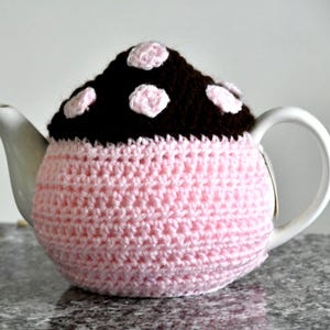 May include: A white ceramic teapot covered with a pink crocheted cozy. The cozy has a dark brown top with light pink polka dots. The teapot has a handle and a spout.
