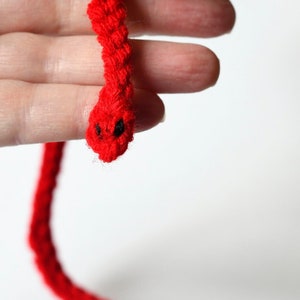 May include: A red crocheted snake with black eyes. The snake is held by a hand.