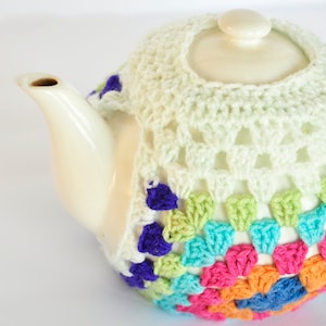 May include: A white ceramic teapot with a cream-colored crocheted cozy. The cozy features a colorful granny square design with pink, orange, blue, green, and purple accents. The teapot has a lid and a handle.