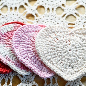 May include: Four crocheted heart-shaped coasters in red, pink, and white. The coasters are arranged in a row on a white crocheted doily.
