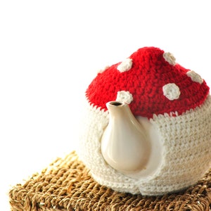 May include: A white ceramic teapot with a red and white crocheted mushroom tea cozy. The cozy has a red top with white polka dots and a white base. The teapot is sitting on a woven basket.