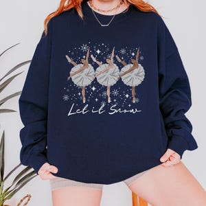 May include: Dark blue sweatshirt with a graphic of three ballerinas in white tutus and pointe shoes, with snowflakes and the text "Let it Snow." The ballerinas have brown skin. A winter-themed design.