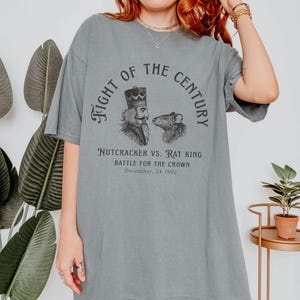 May include: A grey short-sleeve t-shirt featuring a black graphic print of a nutcracker and a rat. The text reads: Fight of the Century, Nutcracker vs. Rat King, Battle for the Crown, December, 24 1892. The shirt has a crew neckline and loose fit.