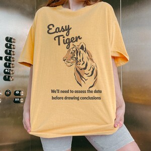 May include: Mustard yellow t-shirt featuring the text "Easy Tiger" and a tiger illustration. The text "We'll need to assess the data before drawing conclusions" is below the tiger. The shirt has a relaxed, oversized style.