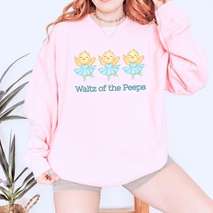 Op de afbeelding: Een lichtroze sweatshirt met drie cartoonkuikens in blauwe tutu's. De tekst "Waltz of the Peeps" staat eronder gedrukt. De sweatshirt is gemaakt van een zacht, comfortabel materiaal.