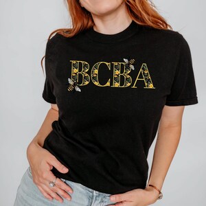 May include: Black t-shirt with the letters "BCBA" in yellow floral design, with two bee illustrations. The shirt is a crew neck style and is being worn by a person.