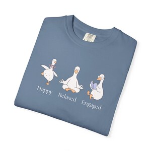 Puede incluir: Camiseta azul con un gráfico de tres gansos blancos en diferentes poses. Uno baila, otro medita y otro lee. Las palabras "Happy", "Relaxed" y "Engaged" están impresas debajo.