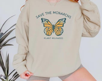 Könnte beinhalten: Beiges Sweatshirt mit langärmligem Monarchfalter-Motiv. Der Schmetterling ist orange und gelb mit schwarzen Details. Der Text "SAVE THE MONARCHS" und "PLANT MILKWEED" ist oberhalb und unterhalb des Schmetterlings aufgedruckt.
