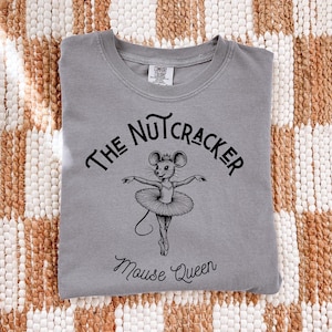 Könnte beinhalten: Ein graues T-Shirt mit Rundhalsausschnitt und einer schwarzen Grafik einer Maus in einem Tutu mit Ballettschuhen. Der Text über der Maus lautet THE NUTCRACKER und der Text darunter Mouse Queen. Das Shirt hat ein Comfort Colors Markenetikett.
