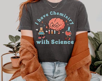 Può includere: T-shirt grigio scuro con la scritta "I have Chemistry with Science" e un cervello a fumetti con gli occhiali. La grafica include attrezzature da laboratorio a fumetti. La persona indossa una giacca marrone e jeans blu.