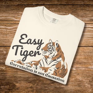 May include: Cream-colored t-shirt with the text "Easy Tiger" in black script and a brown and black tiger illustration. The phrase "Correlation is not Causation" is printed below the tiger. The shirt is folded on a wooden surface.