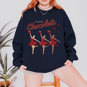 May include: Navy blue sweatshirt with a graphic of three ballerinas in red dresses holding chocolate boxes. The word "Chocolate" is in red script above the dancers, with the word "Nutcracker" above it.