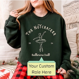May include: A dark green crewneck sweatshirt featuring a cream-colored line art graphic of a ballerina. The text reads THE NUTCRACKER, 20 26, and Ballerina Doll. A light green speech bubble overlay at the bottom displays the text Your Custom Role Here.