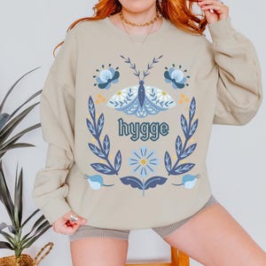 May include: Beige sweatshirt with a floral and butterfly design. The design includes blue flowers, a blue and white butterfly, and the word "hygge" in green. The sweatshirt has long sleeves and a crew neck.