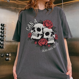 以下が含まれることがあります： チャコールグレーのTシャツで、赤いバラと緑の葉に囲まれた2つの頭蓋骨のグラフィックプリントが施されています。頭蓋骨の上には「CAN A」という文字がアーチ状に配置され、下には「DEAD MAN DO IT?」という文字が曲線を描いてプリントされています。