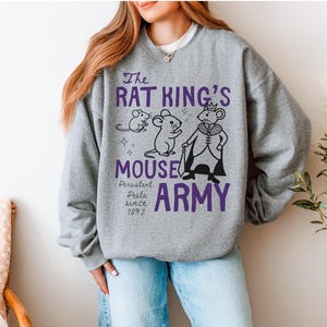 May include: A heather gray crewneck sweatshirt featuring a graphic print in purple and black. The design includes three mice, one wearing a crown and cape, with the text "The RAT KING'S MOUSE ARMY" and "Persistent Pests since 1892" printed on the front.