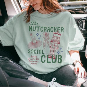 May include: A light green t-shirt featuring a graphic print with the text The Nutcracker Social Club and Sorry I can't. It's Nutcracker season. The design includes a red tutu, ballet slippers, and a nutcracker figure in red and blue ink.