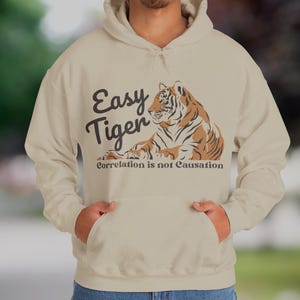 May include: Beige hoodie with a tiger graphic. The design features a tiger illustration in shades of brown and orange, with the words "Easy Tiger" and "Correlation is not Causation" in black text.