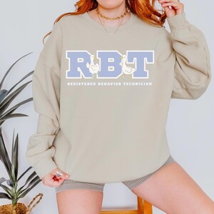 May include: A beige sweatshirt with the letters "RBT" in light blue, with two cartoon ducks. Below the letters, the text reads "REGISTERED BEHAVIOR TECHNICIAN". The sweatshirt has long sleeves and a crew neck.